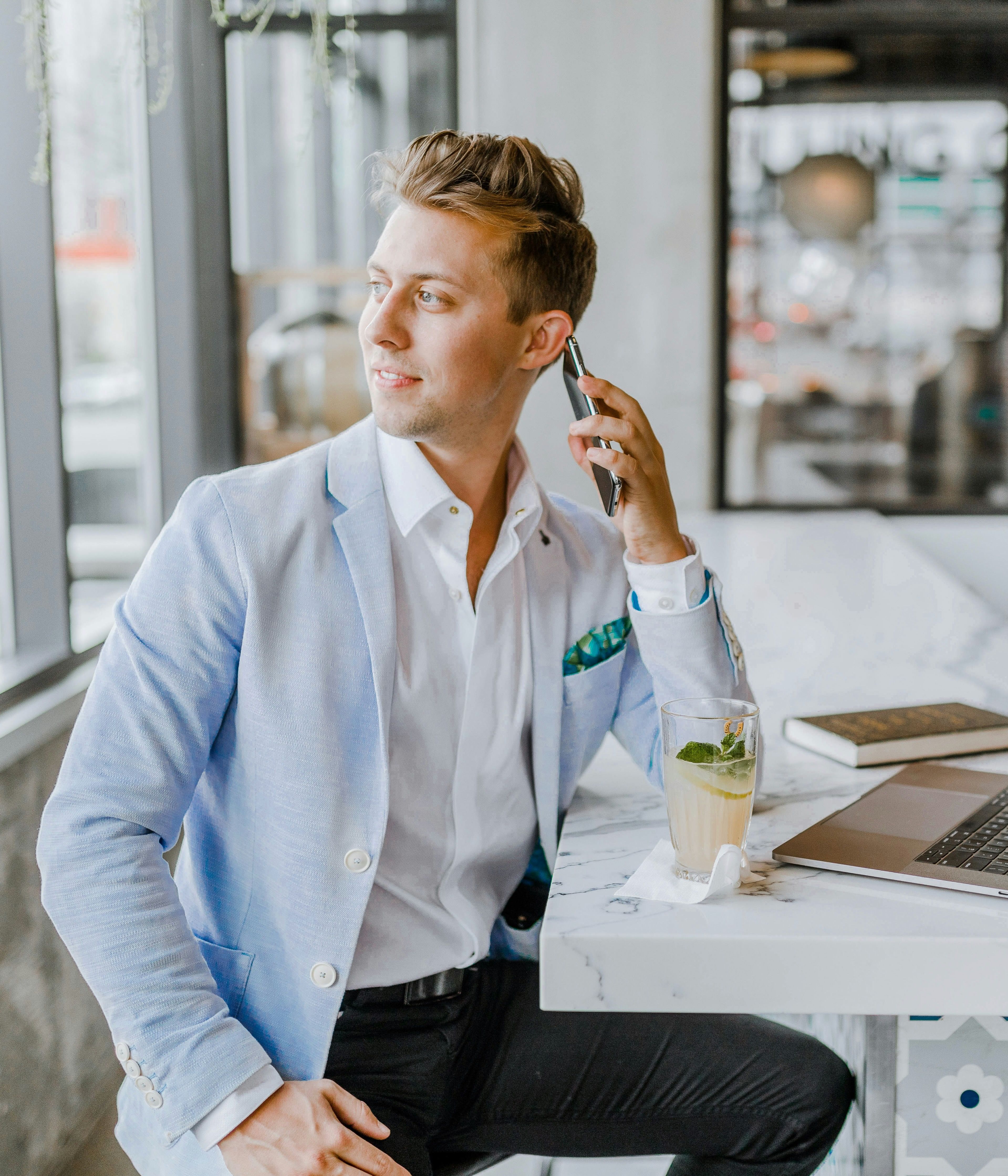 A young professional in a light blue blazer sitting at a cafe bar, holding a phone to his ear, with a laptop and a drink on the counter.